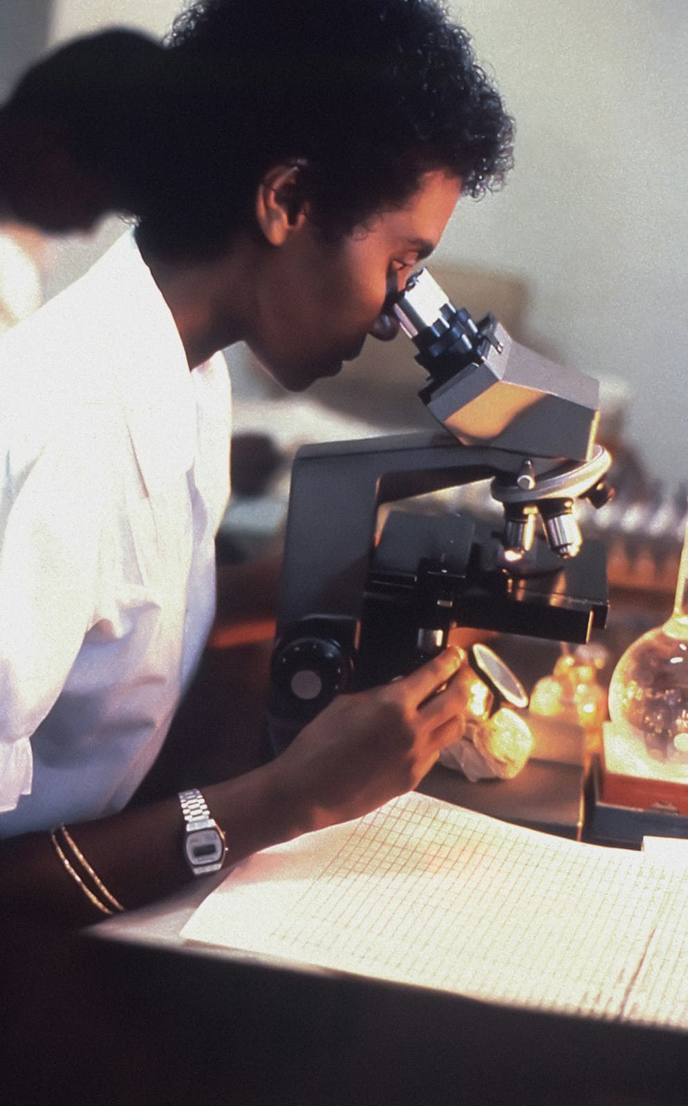 A researcher at a microscope, with notes on graph paper under warm lamp light